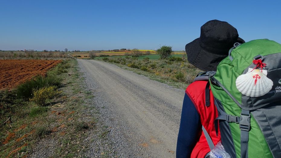 A pilgrim walking along the gravel path on the Camino de Santiago, with a backpack adorned with the iconic scallop shell symbol